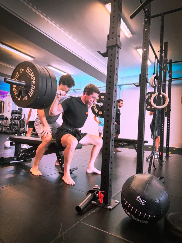 Adult performing a barbell back squat with a coach spotting, at Be Water Lisboa